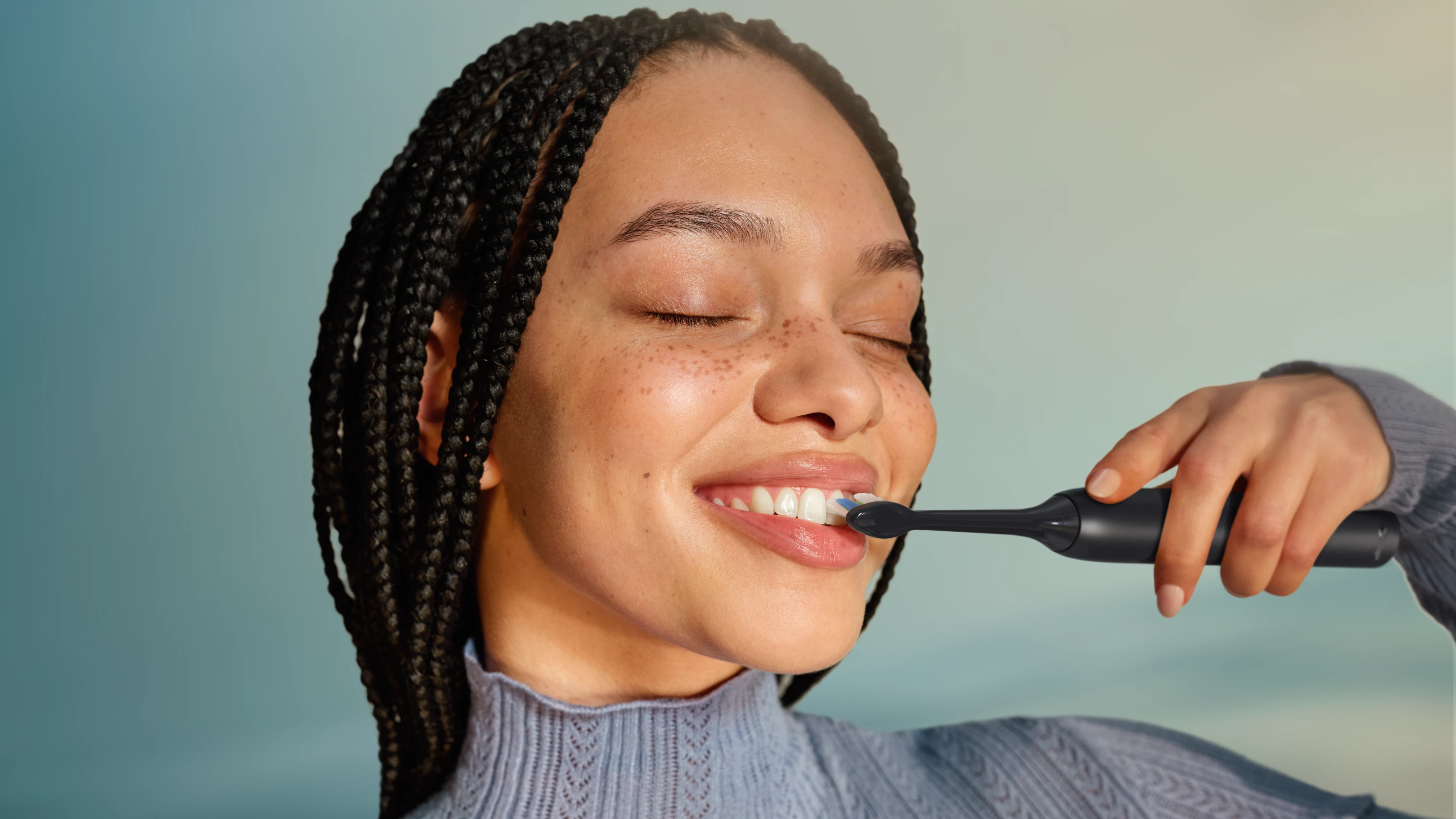 Kobieta z czarnymi warkoczami myje zęby czarną szczoteczką elektryczną.