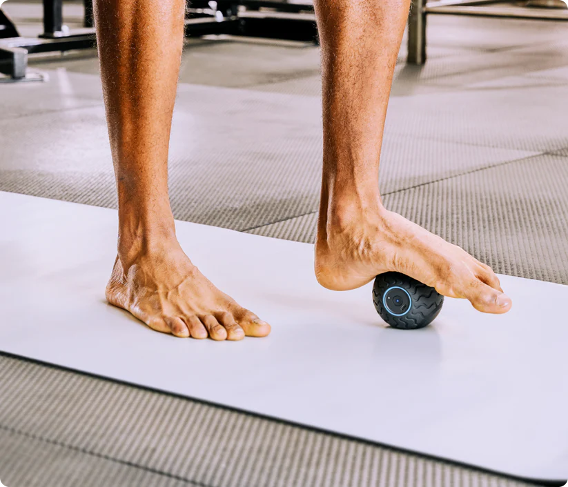 Un hombre usa una pelota de masaje negra en su pie, sobre una colchoneta en un gimnasio.