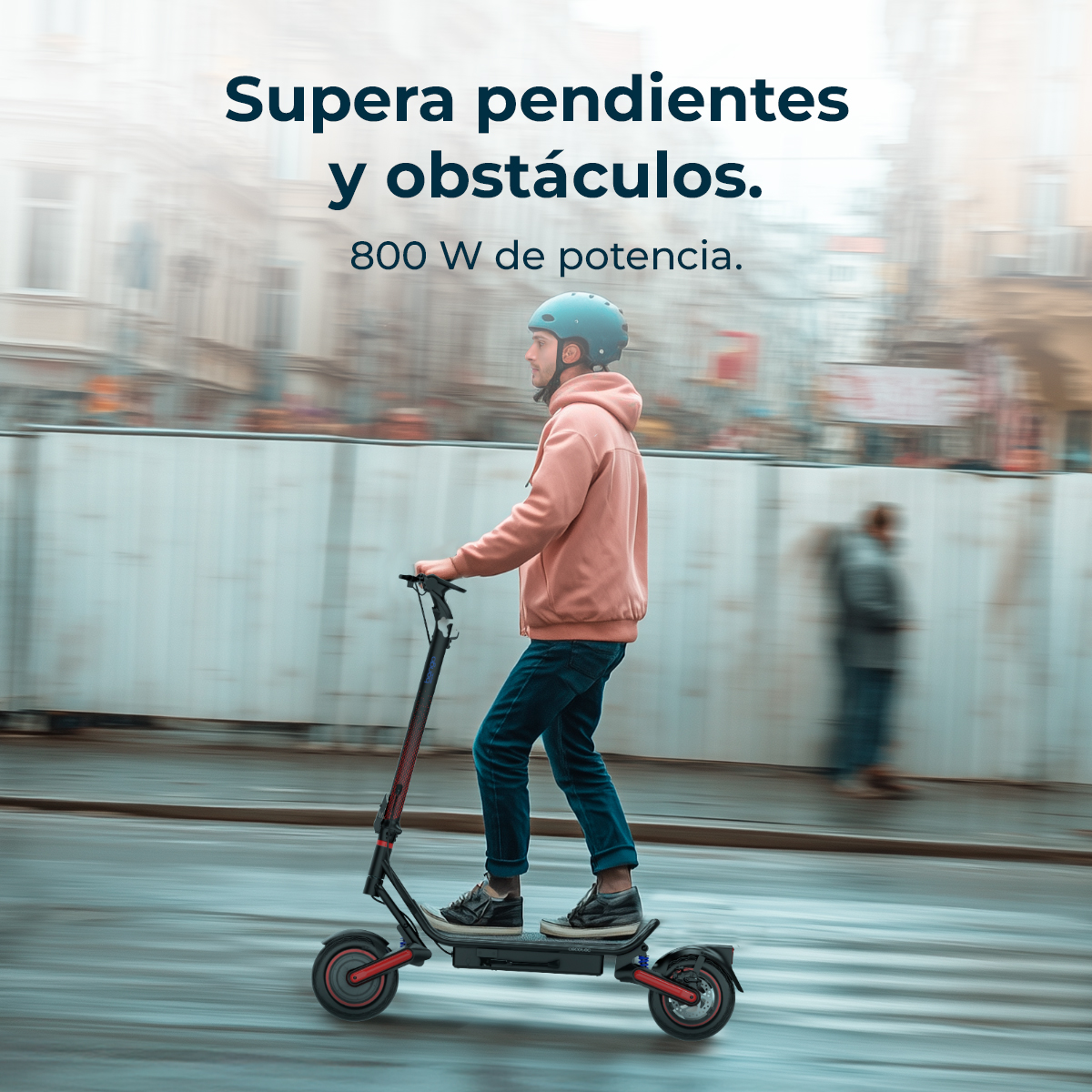 Hombre monta patinete eléctrico con casco, sudadera rosa, jeans azules, en calle mojada.