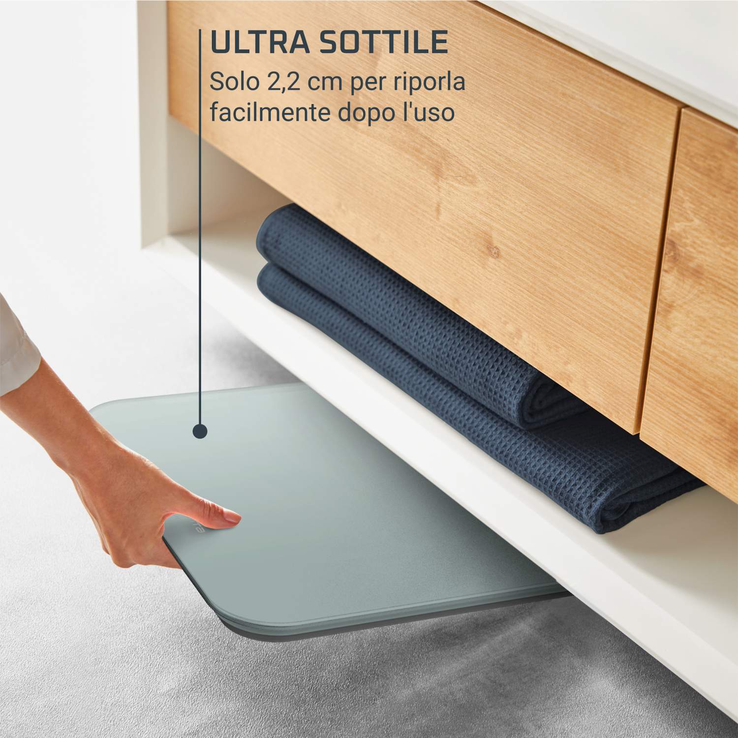 Una mano estrae una bilancia sottile e grigia da sotto una mensola di legno con asciugamani scuri.