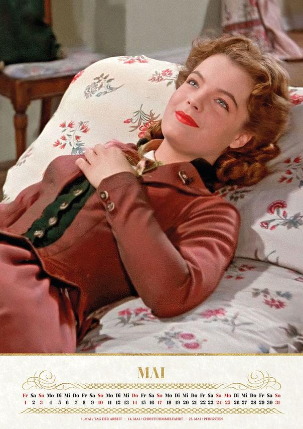 Eine Frau mit roten Lippen liegt auf einem gemusterten Sofa und trägt eine braune Jacke.
