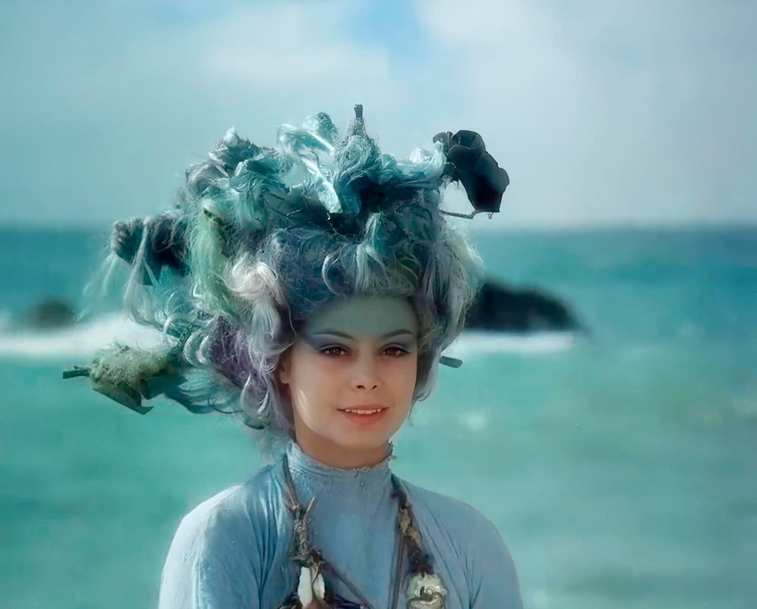Eine Frau mit blaugrünem Gesicht und fantasievollem Haar steht am Meer.