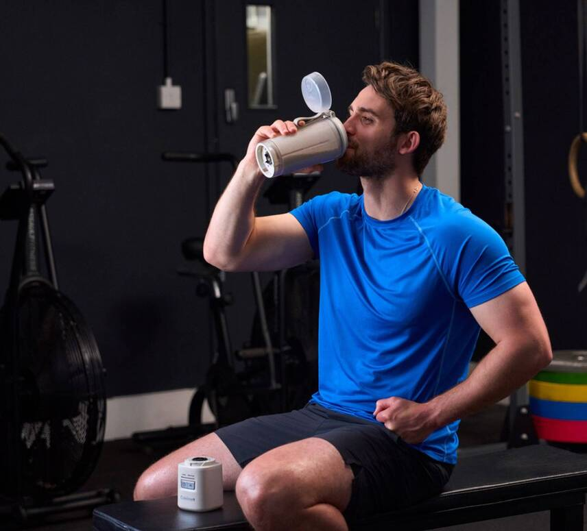 Homme en chemise bleue boit dans un shaker au gym, vélo à air derrière lui.