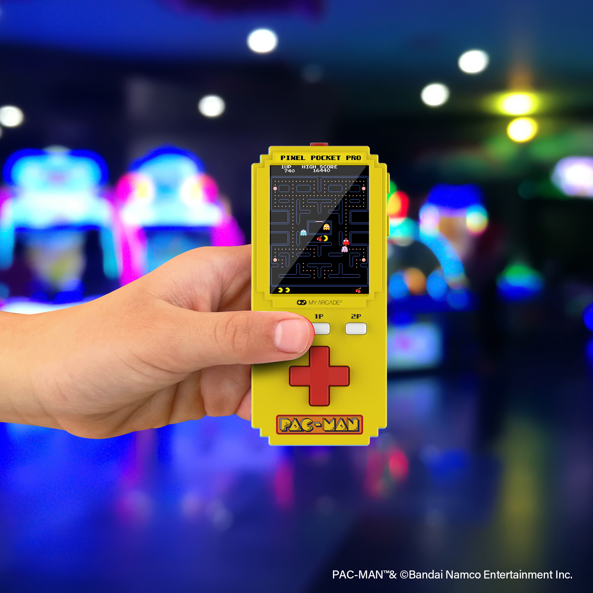 Gelbes Pac-Man Handheld-Spielgerät wird in einer Spielhalle gehalten. Bunte Lichter im Hintergrund.