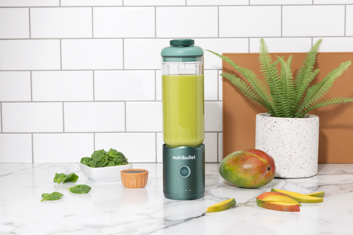Una licuadora verde con un batido verde se encuentra junto a mangos y espinacas.