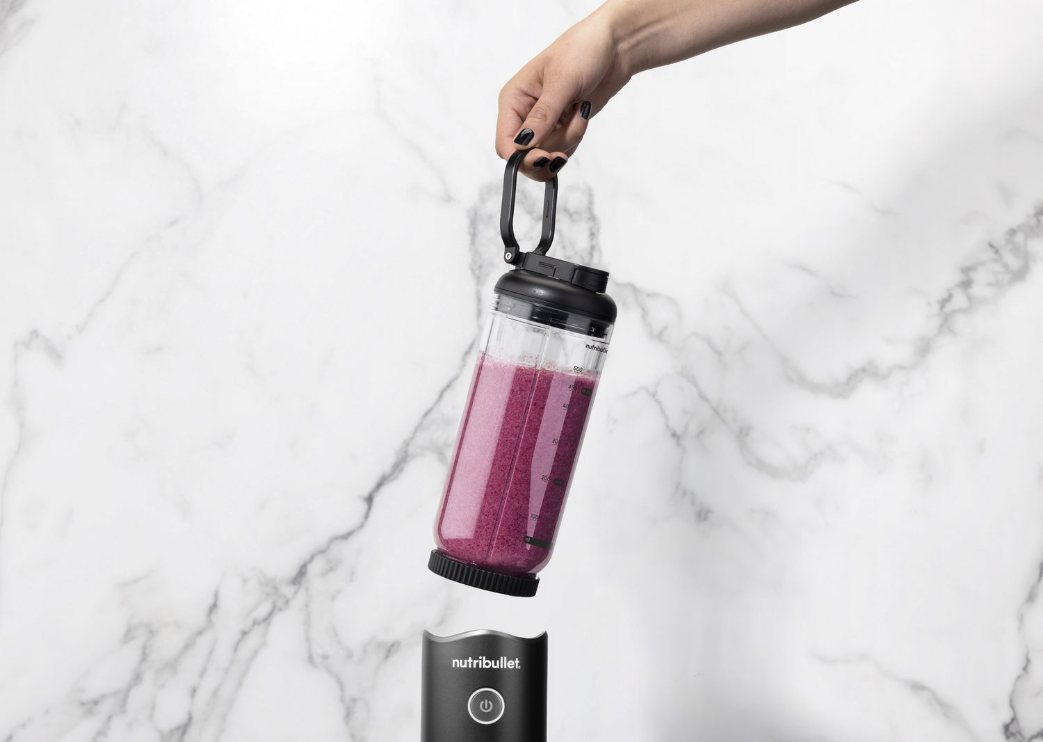 Mano sostiene botella licuadora con batido morado sobre base en fondo mármol.