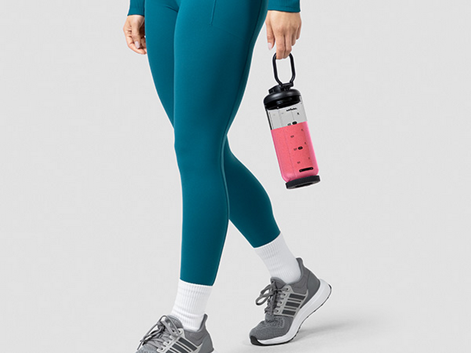 Mujer con ropa deportiva turquesa sostiene una licuadora portátil con líquido rosa.