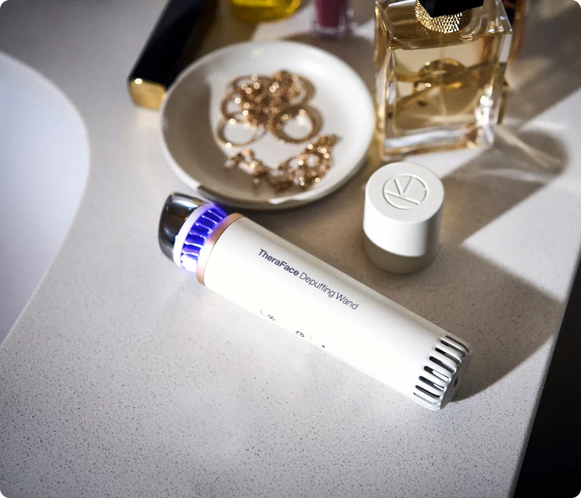 Una varita Theragun blanca con luz azul descansa sobre un mostrador cerca de joyas y perfume.