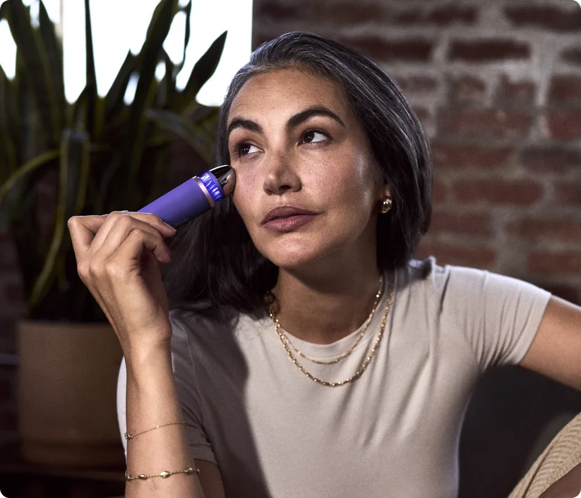 Una mujer con cabello entrecano usa un dispositivo púrpura para el cuidado de la piel.
