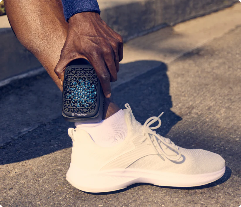 Dispositivo Theragun negro en el tobillo de una persona con zapato blanco.