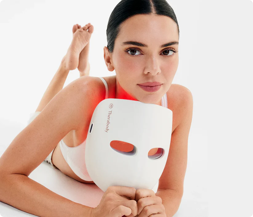 Mujer con mascarilla Therabody y luces rojas, fondo blanco.
