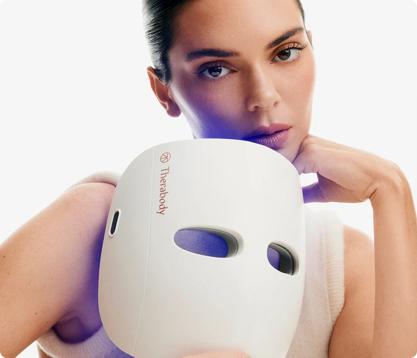 Una mujer sostiene una mascarilla Therabody blanca con luces azules frente a su rostro.