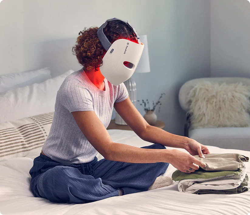 Una mujer se sienta en una cama doblando ropa mientras usa una mascarilla facial de luz roja.