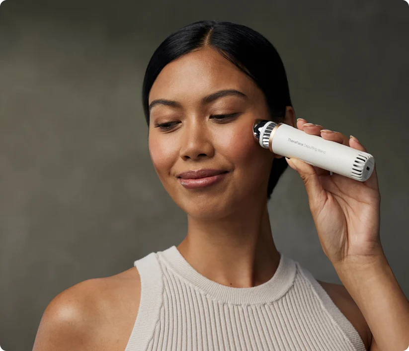 Una mujer de cabello oscuro y top de canalé crema usa un dispositivo facial blanco en su mejilla derecha, sonriendo suavemente.