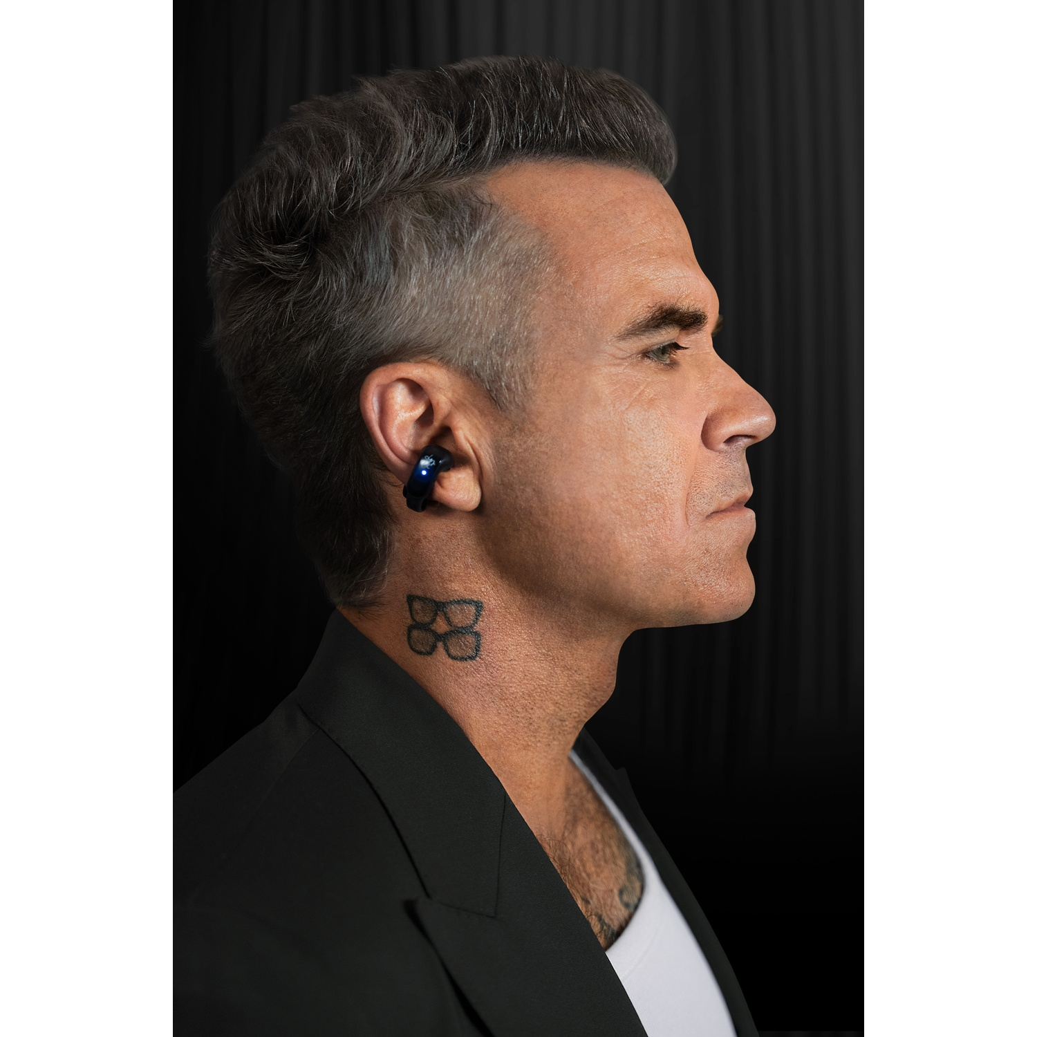 Man with an earbud and glasses tattoo in profile against a dark background.