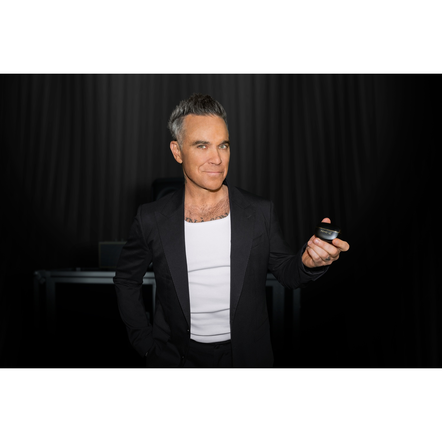 A man in a black suit and white tank top holds a small black device.