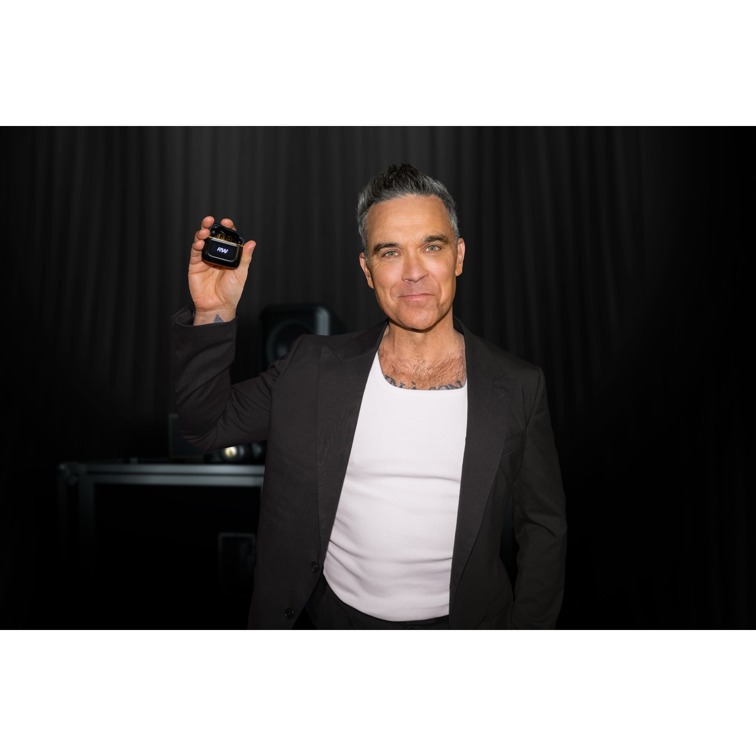 Man in black suit and white tank top holds earbud charging case.