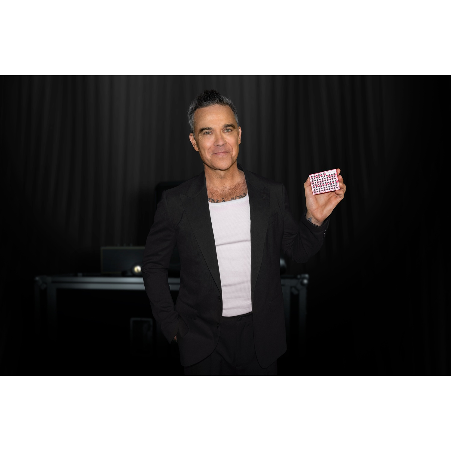 Robbie Williams in a black suit holds a small box with red dots.