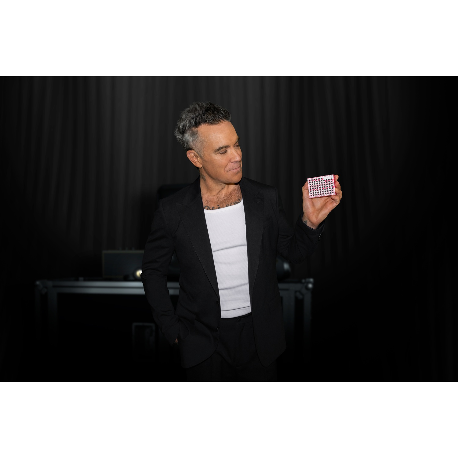 Man in a black suit holds a small red box with white dots.