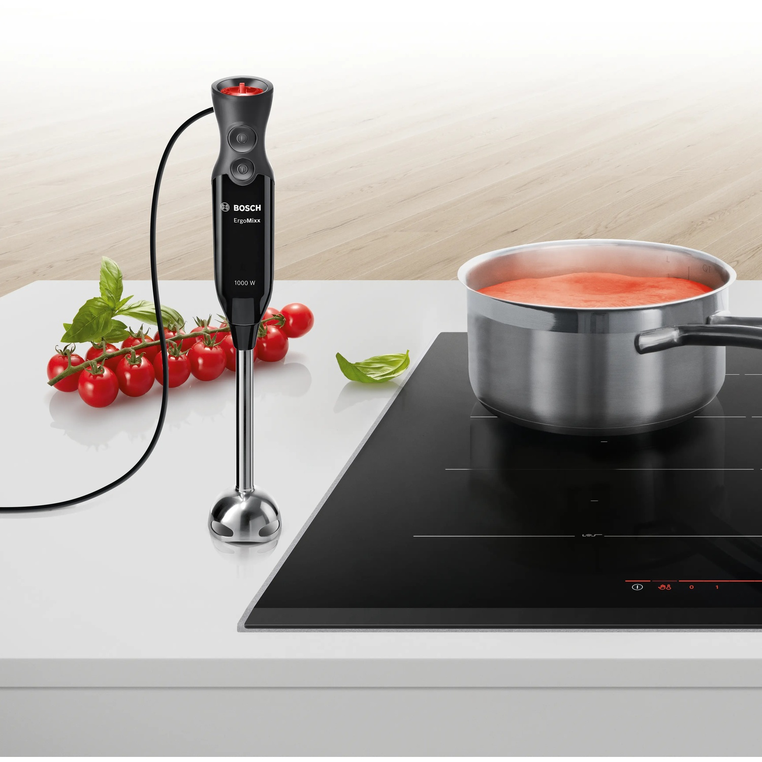 A black immersion blender with tomatoes and a pot on an induction cooktop.