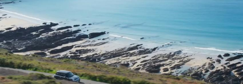 Ein dunkles Auto fährt auf einer Küstenstraße mit Blick auf einen felsigen Strand und das Meer.