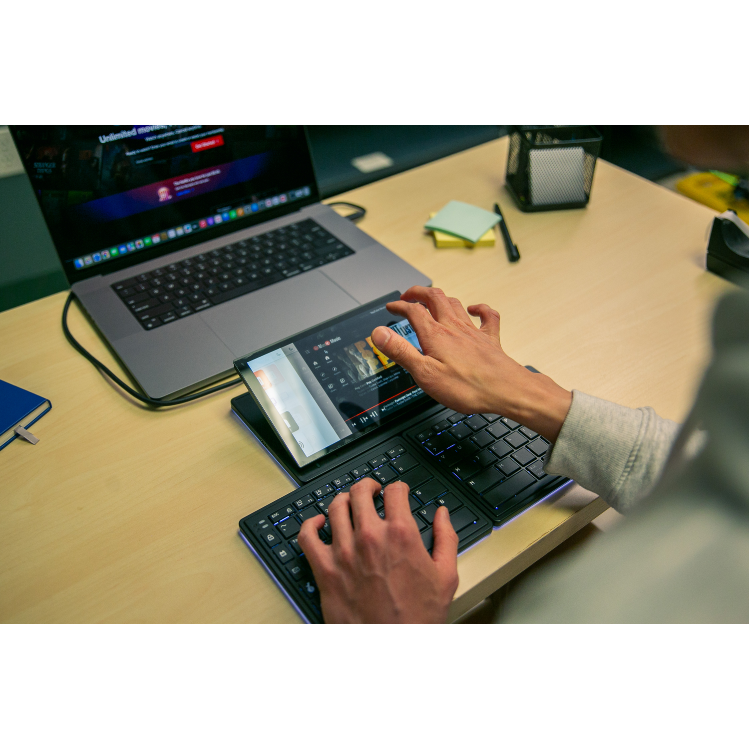 Hands operating a tablet and keyboard next to a laptop on a desk.