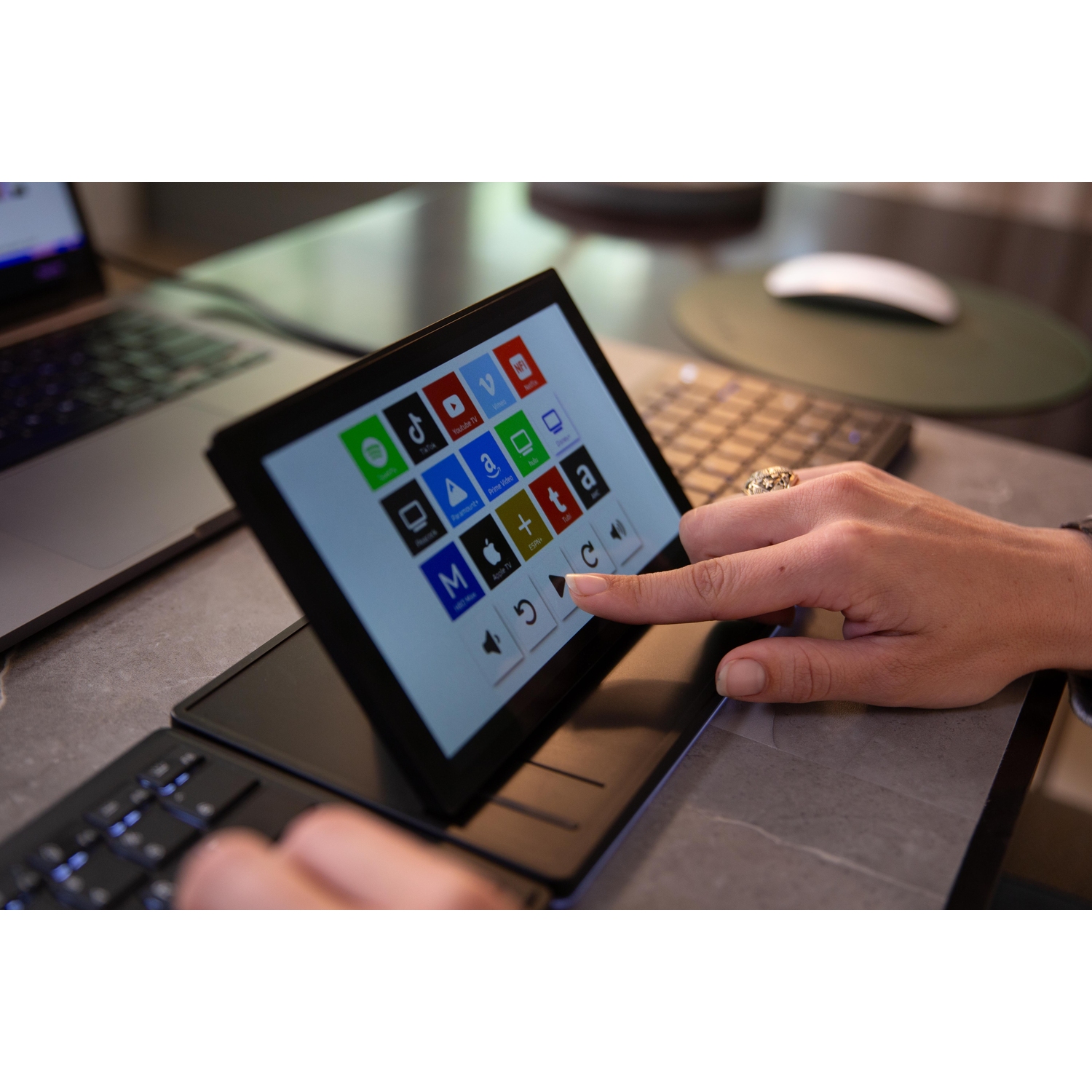 A hand taps a small screen displaying app icons. A laptop is in the background.