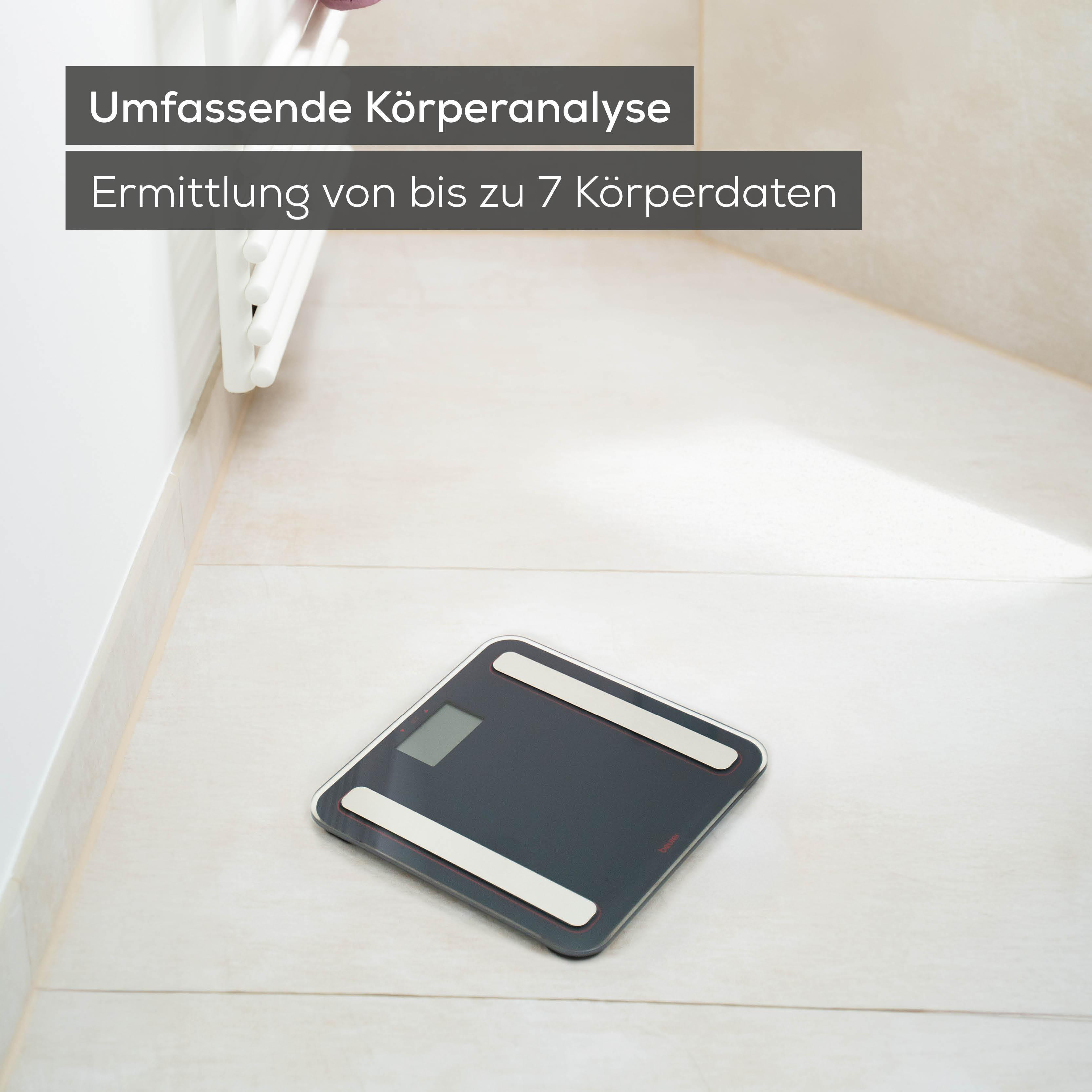 Eine dunkle Körperwaage mit zwei weißen Streifen und einem Display steht auf hellem Fliesenboden.