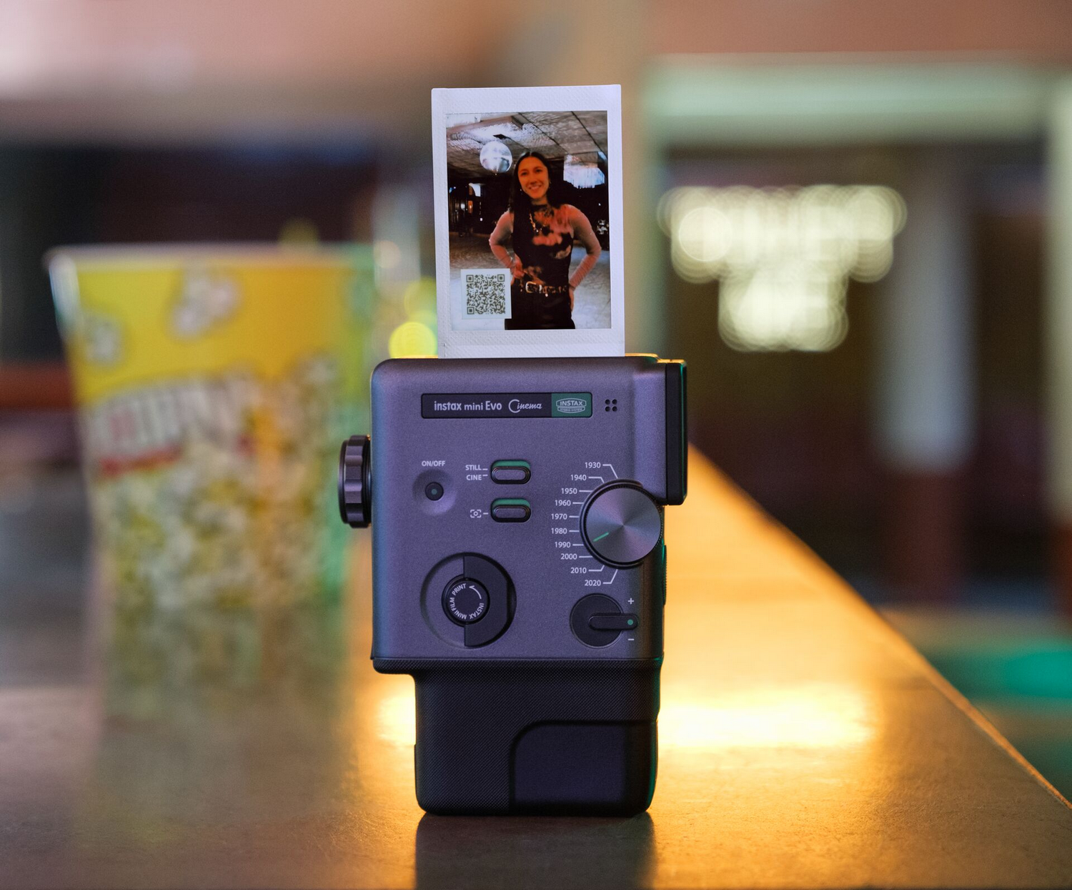 Cámara Instax Mini Evo con foto y cubo de palomitas de fondo.