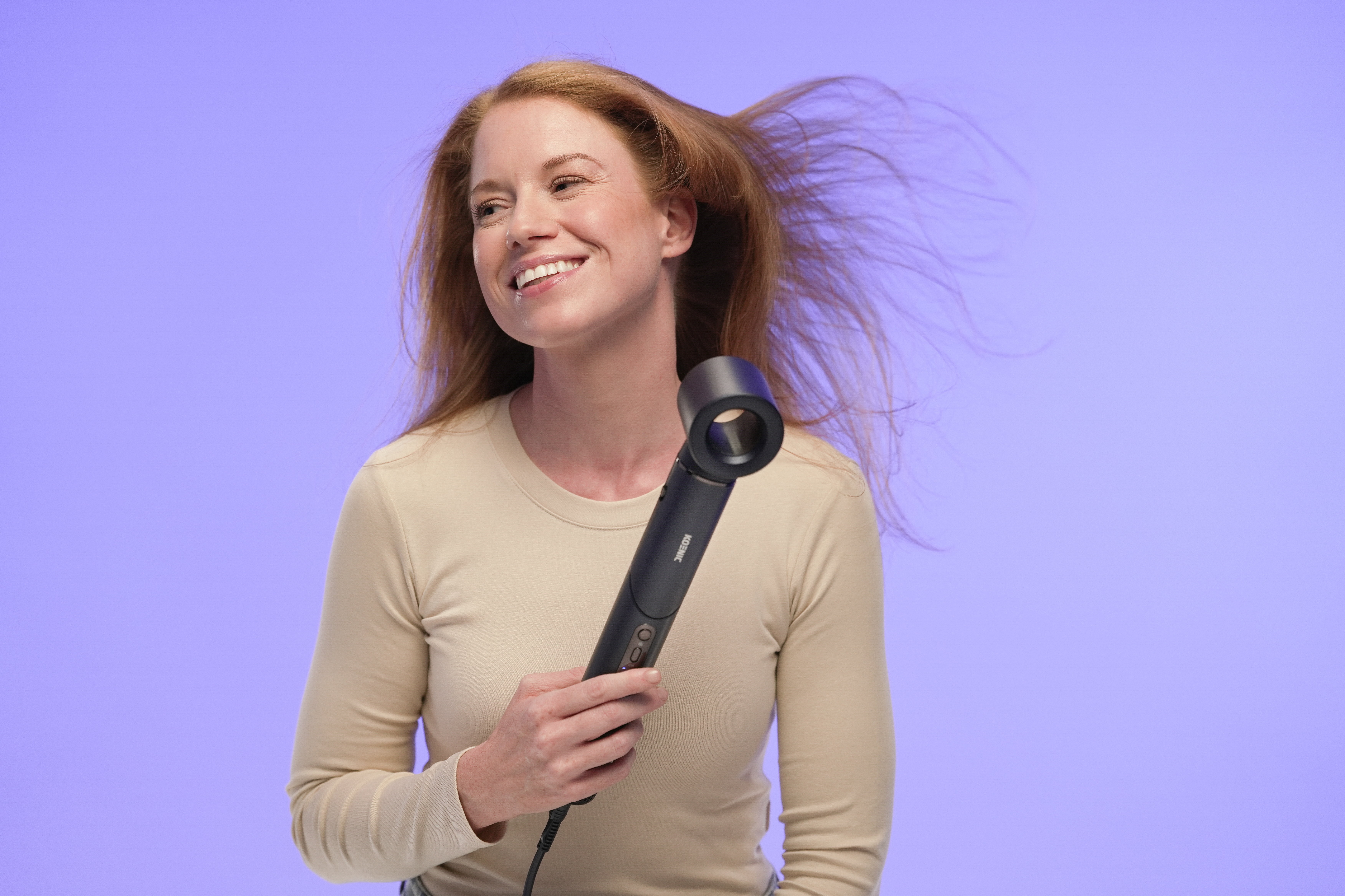 Kobieta z rudymi włosami używa czarnej suszarki do włosów na fioletowym tle.