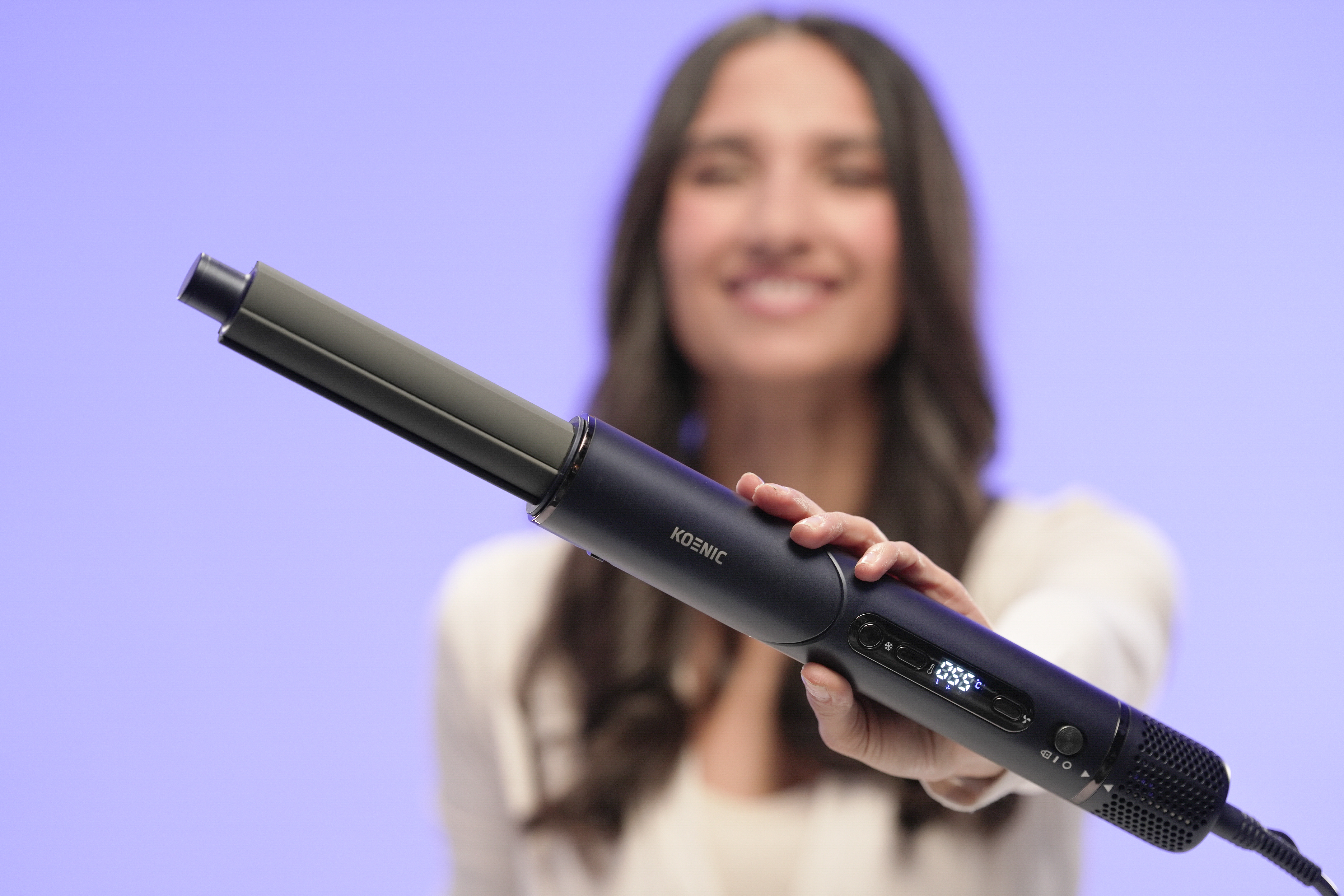 Kobieta trzyma ciemną lokówkę z cyfrowym wyświetlaczem na fioletowym tle.