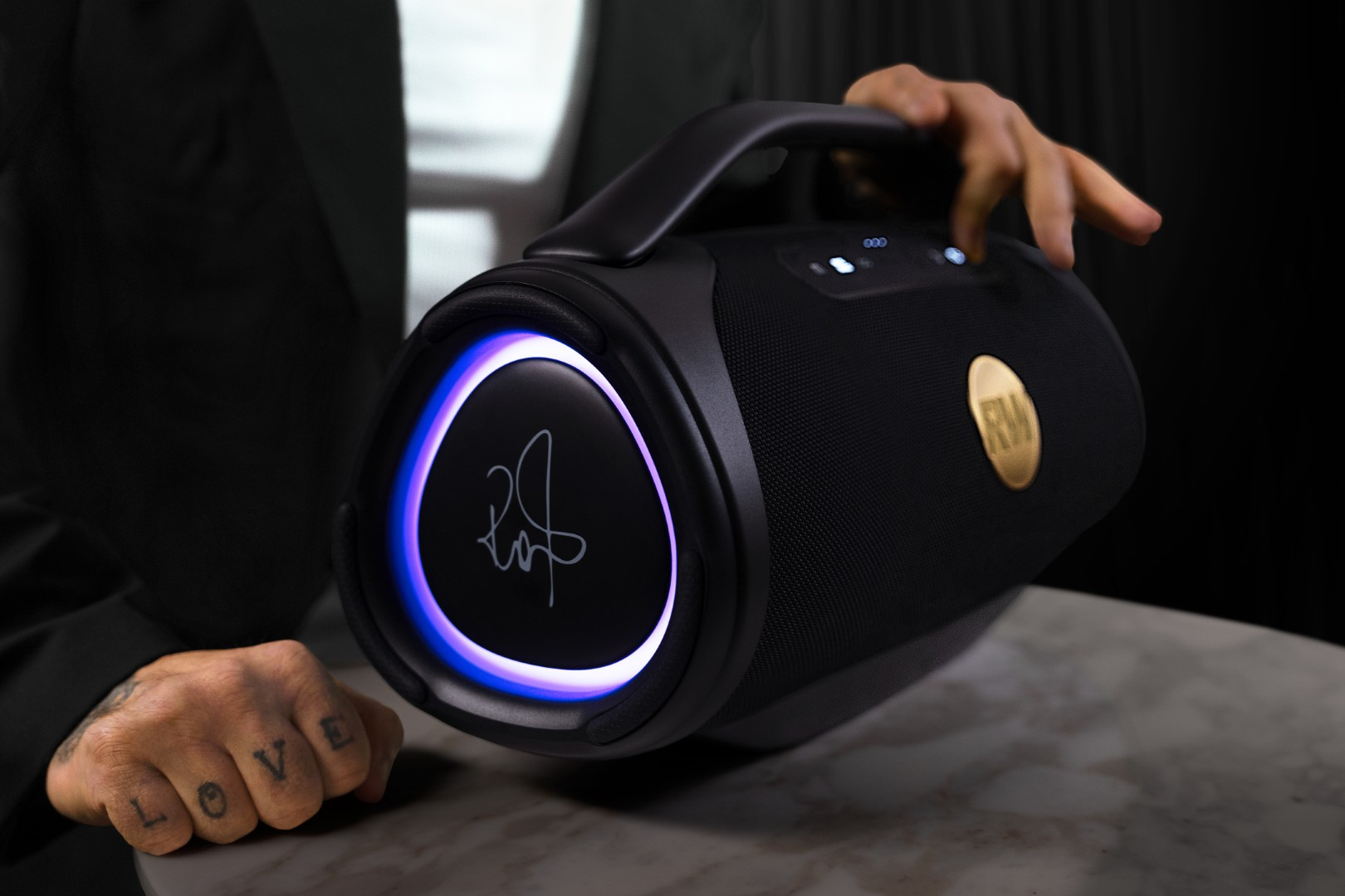 Czarny głośnik z niebieskim pierścieniem świetlnym, złotym logo i uchwytem. Ręka z tatuażem.
