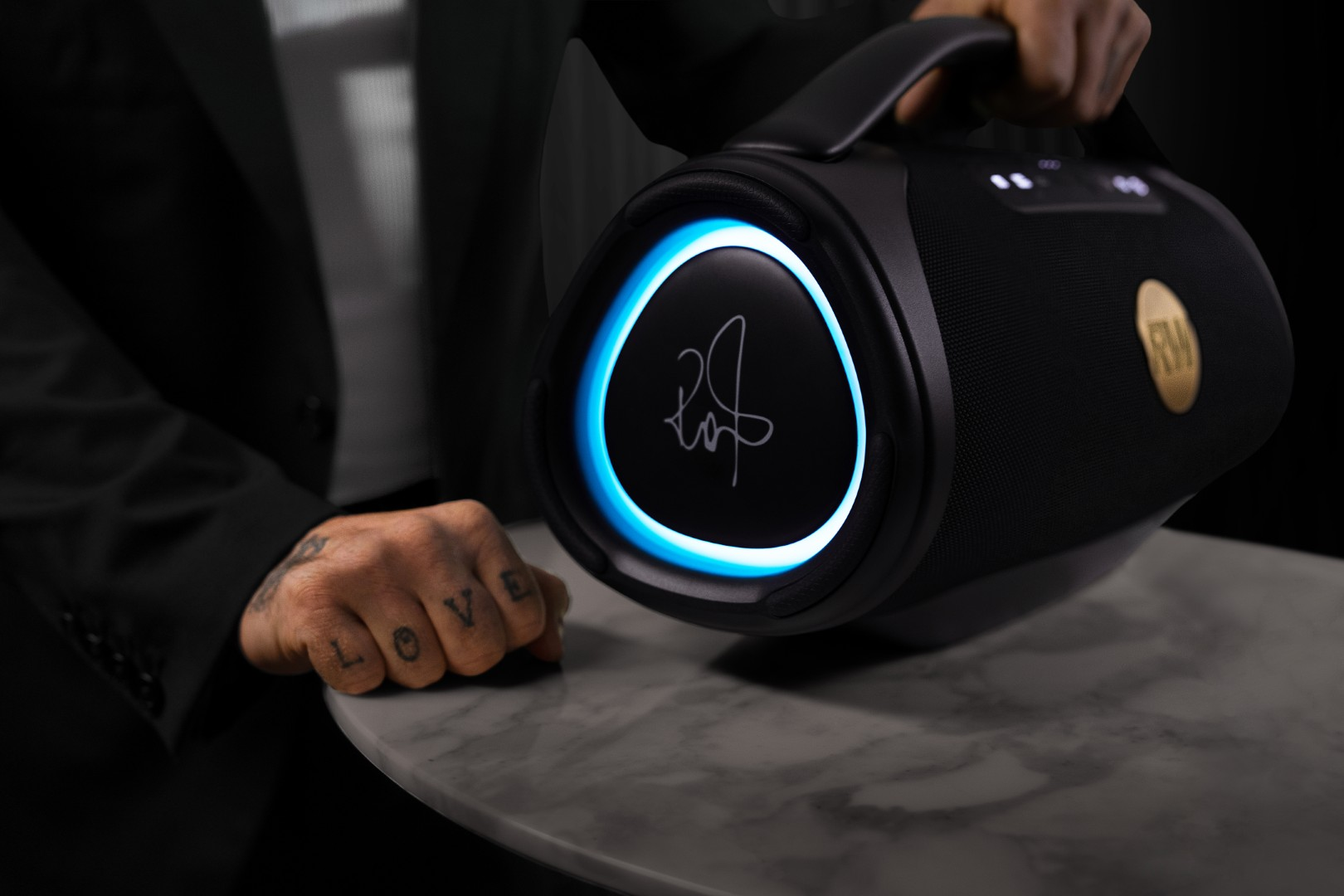 Czarny głośnik z niebieskim pierścieniem świetlnym i uchwytem trzymany przez osobę.