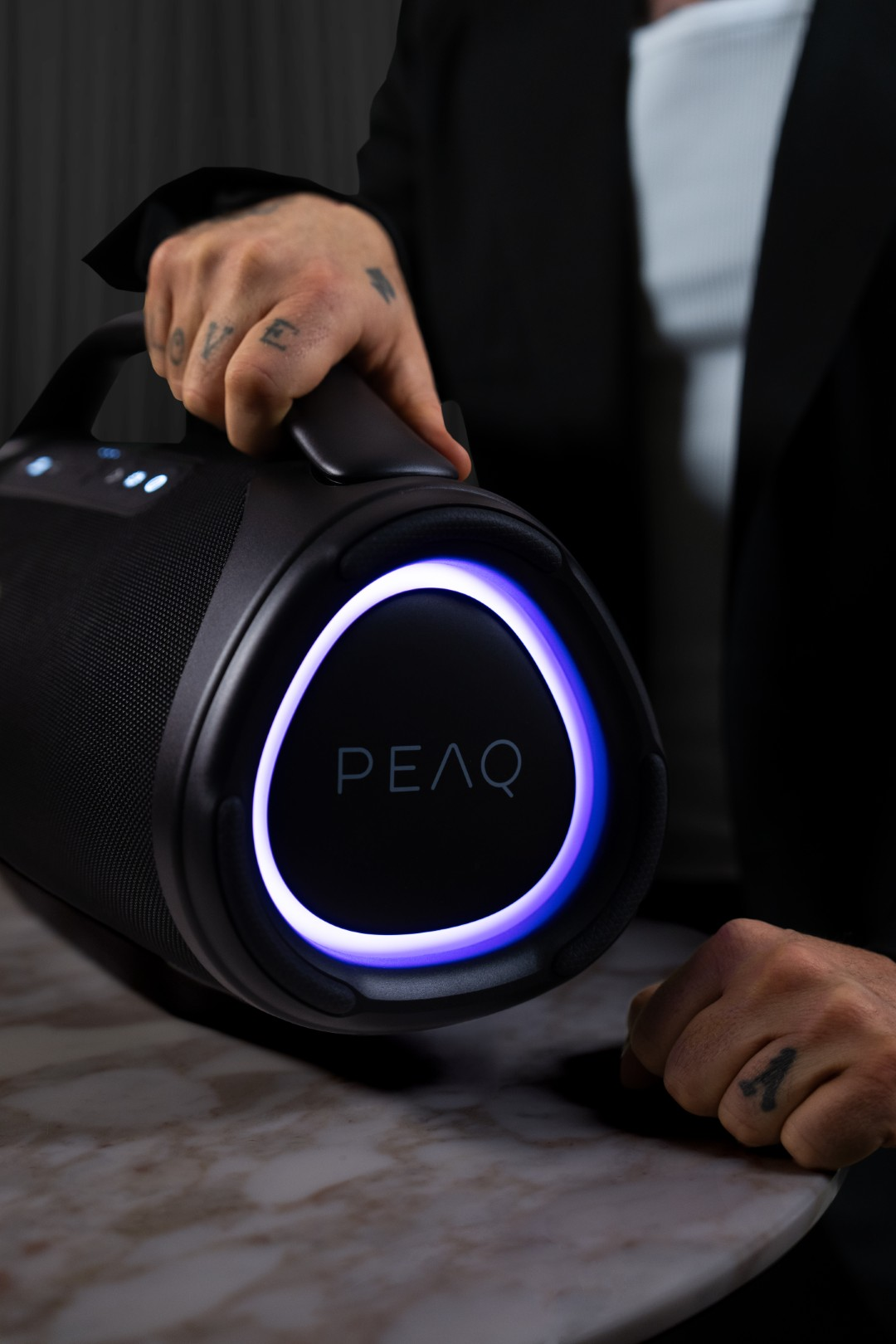 Czarny głośnik z niebieskim pierścieniem świetlnym i logo "PEAQ", trzymany przez osobę.