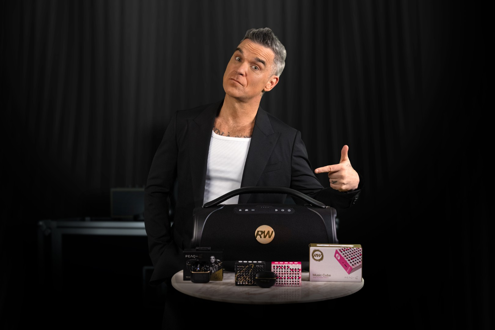 Robbie Williams wskazuje na czarną boombox i kolorowe kostki muzyczne na stole.