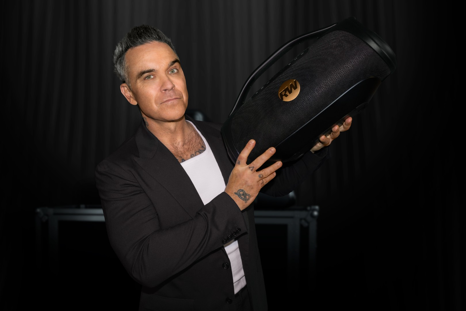 Mężczyzna z siwiejącymi włosami w czarnym garniturze trzyma czarny głośnik ze złotym logo.