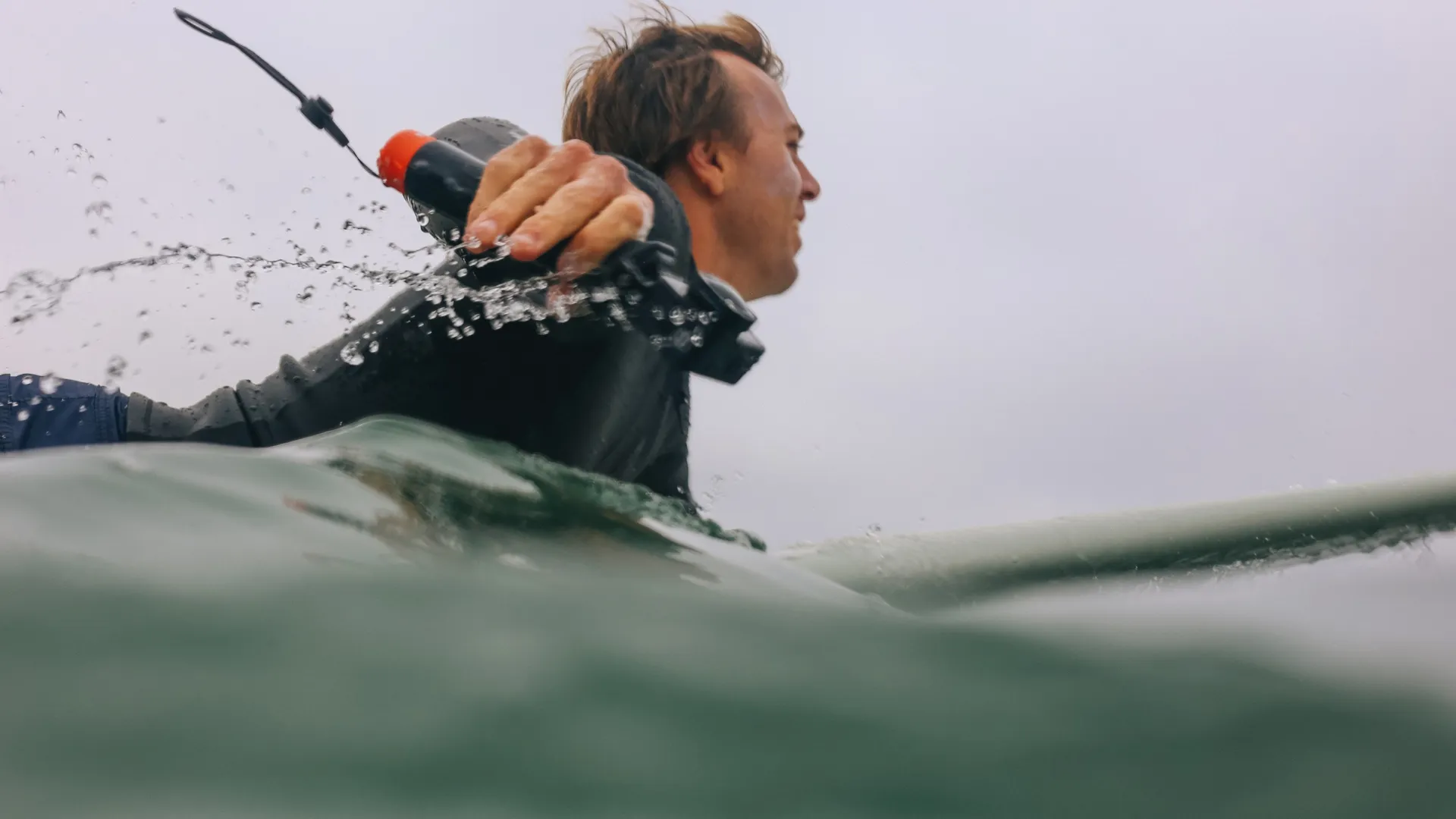 Ein Mann in einem Neoprenanzug ist von der Brust aufwärts zu sehen, wie er sich auf einem Surfbrett durch grünes Wasser unter einem grauen Himmel bewegt.