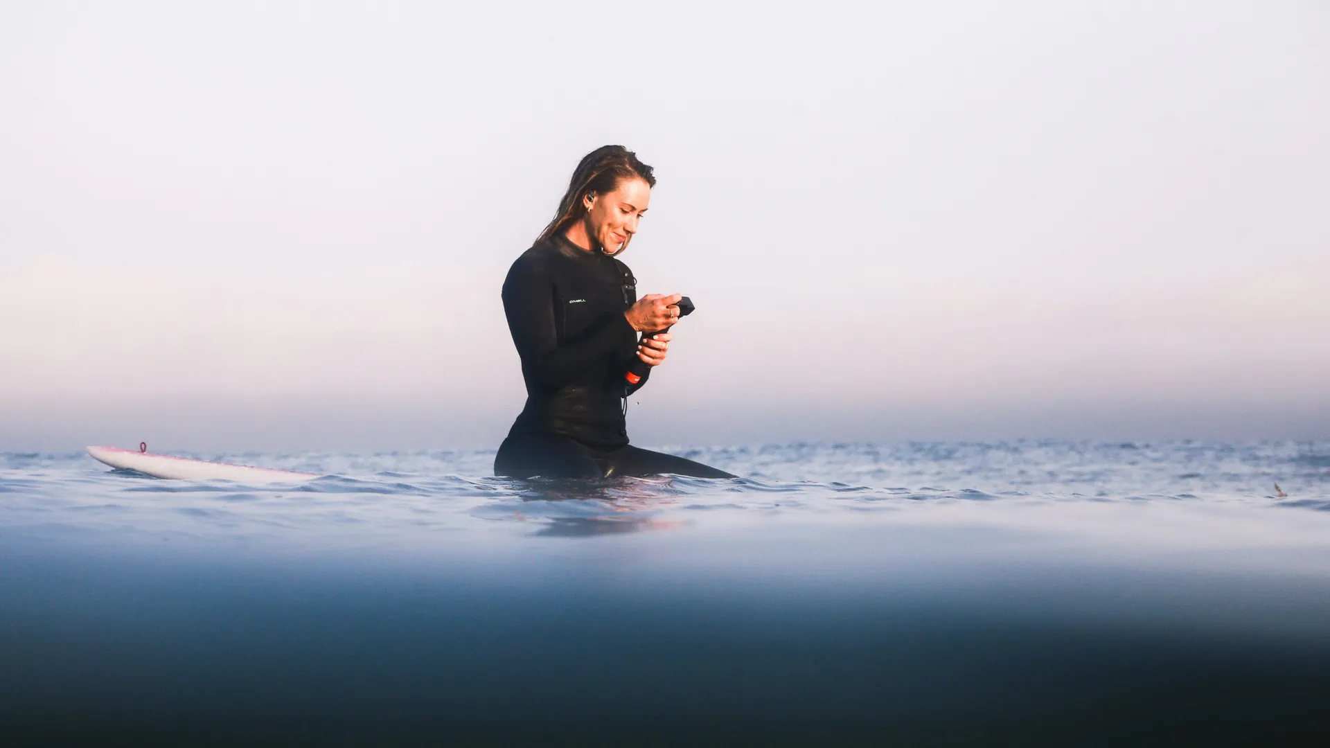 Frau im schwarzen Neoprenanzug sitzt auf Surfbrett im Meer und schaut auf ihr Handy.