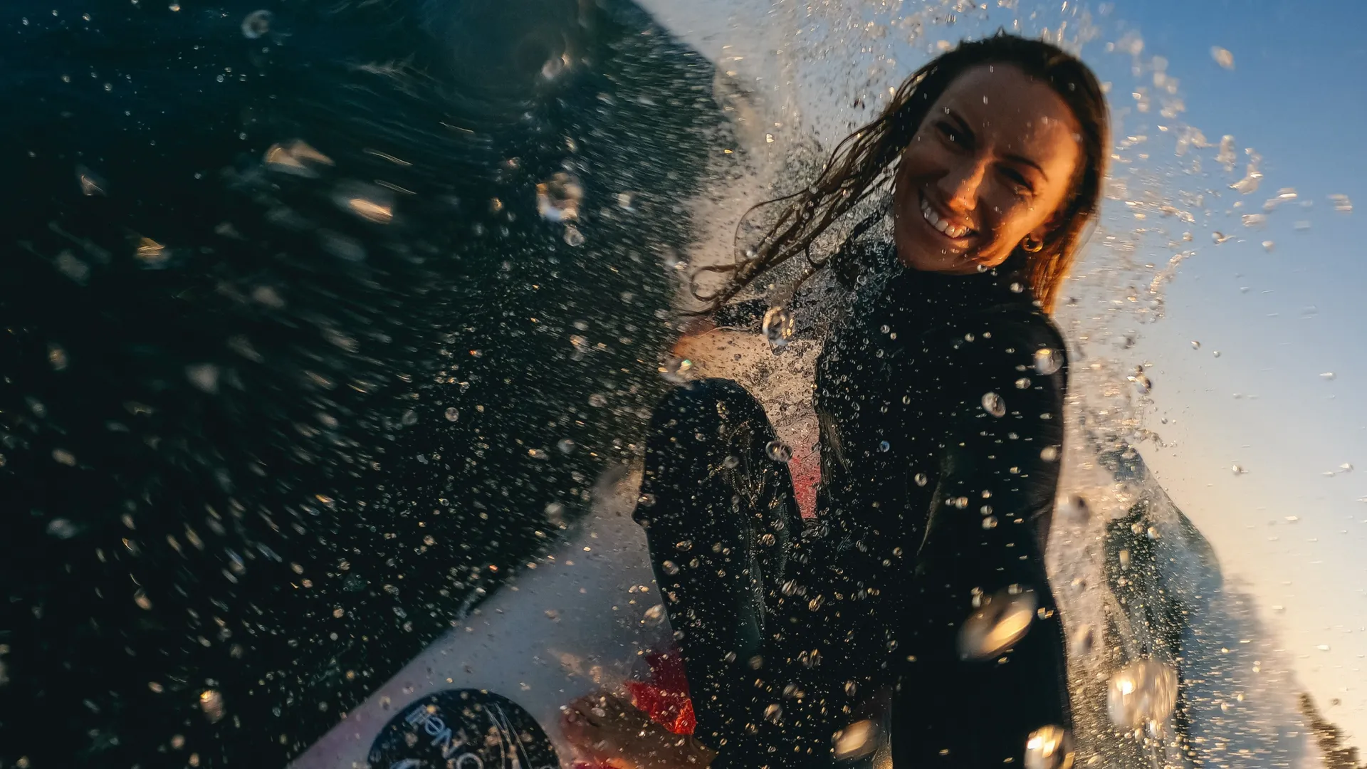 Eine lachende Surferin auf einem Surfbrett, umgeben von Wasserspritzern.
