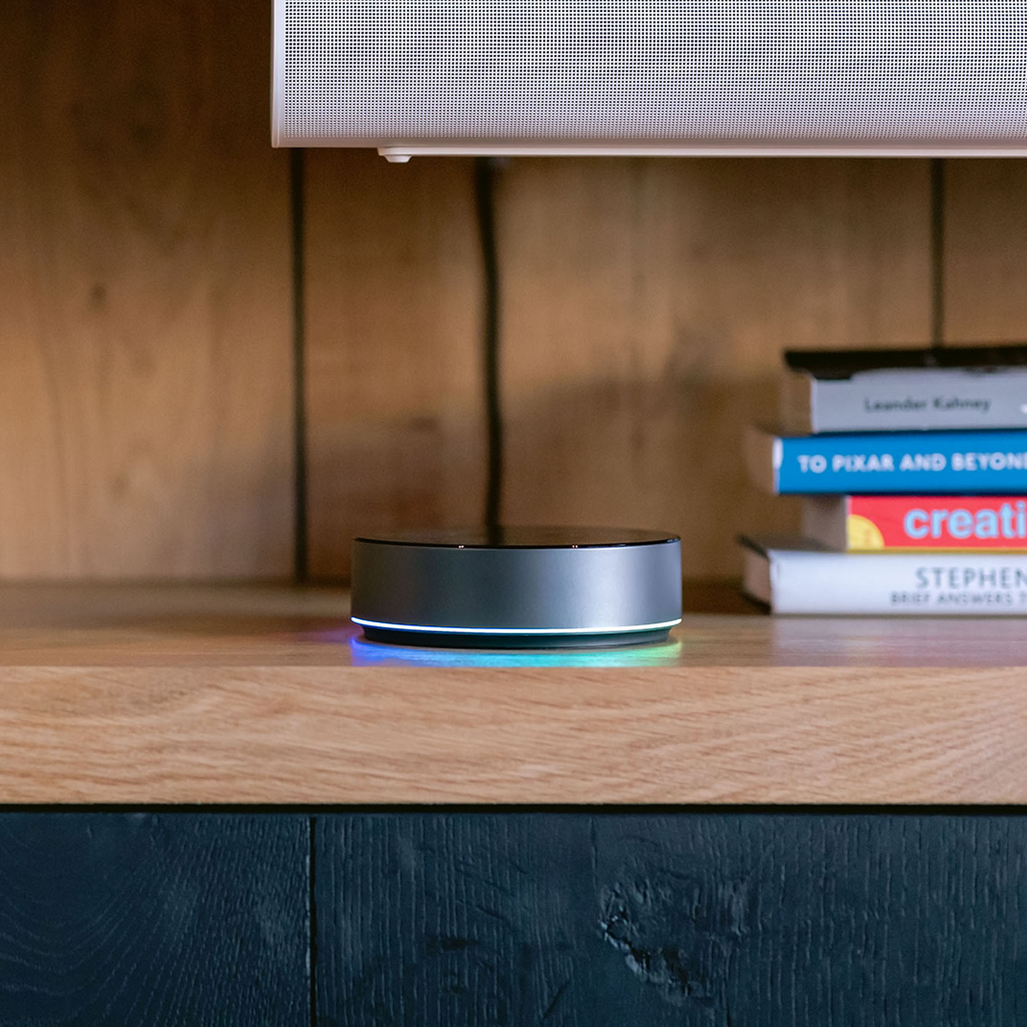 Un altavoz inteligente plateado con luz azul en un estante de madera junto a libros.