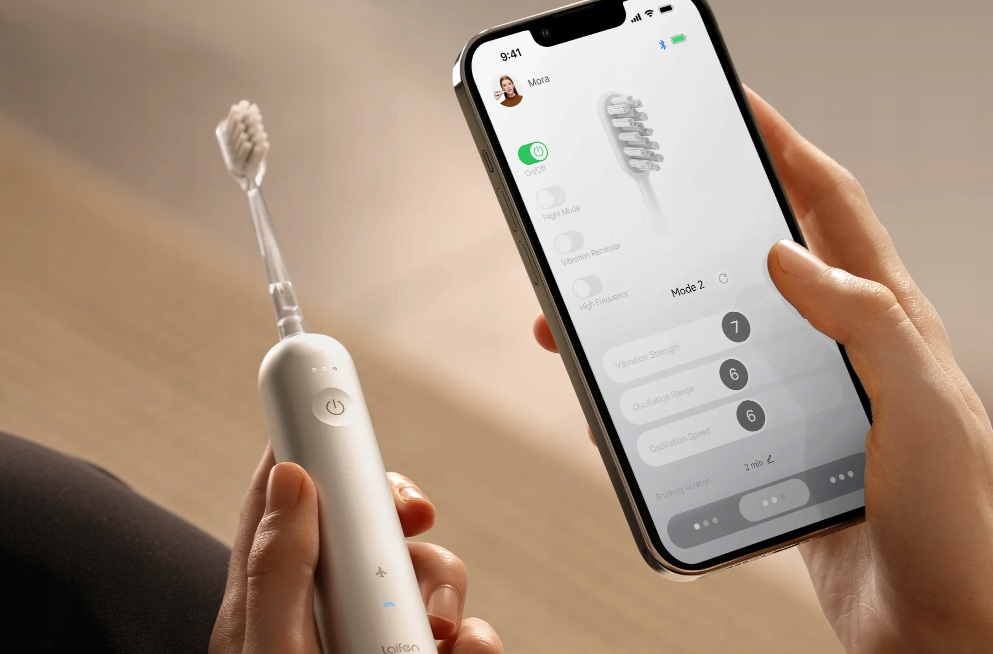 Osoba trzyma białą szczoteczkę elektryczną i smartfon z aplikacją sterującą szczoteczką.