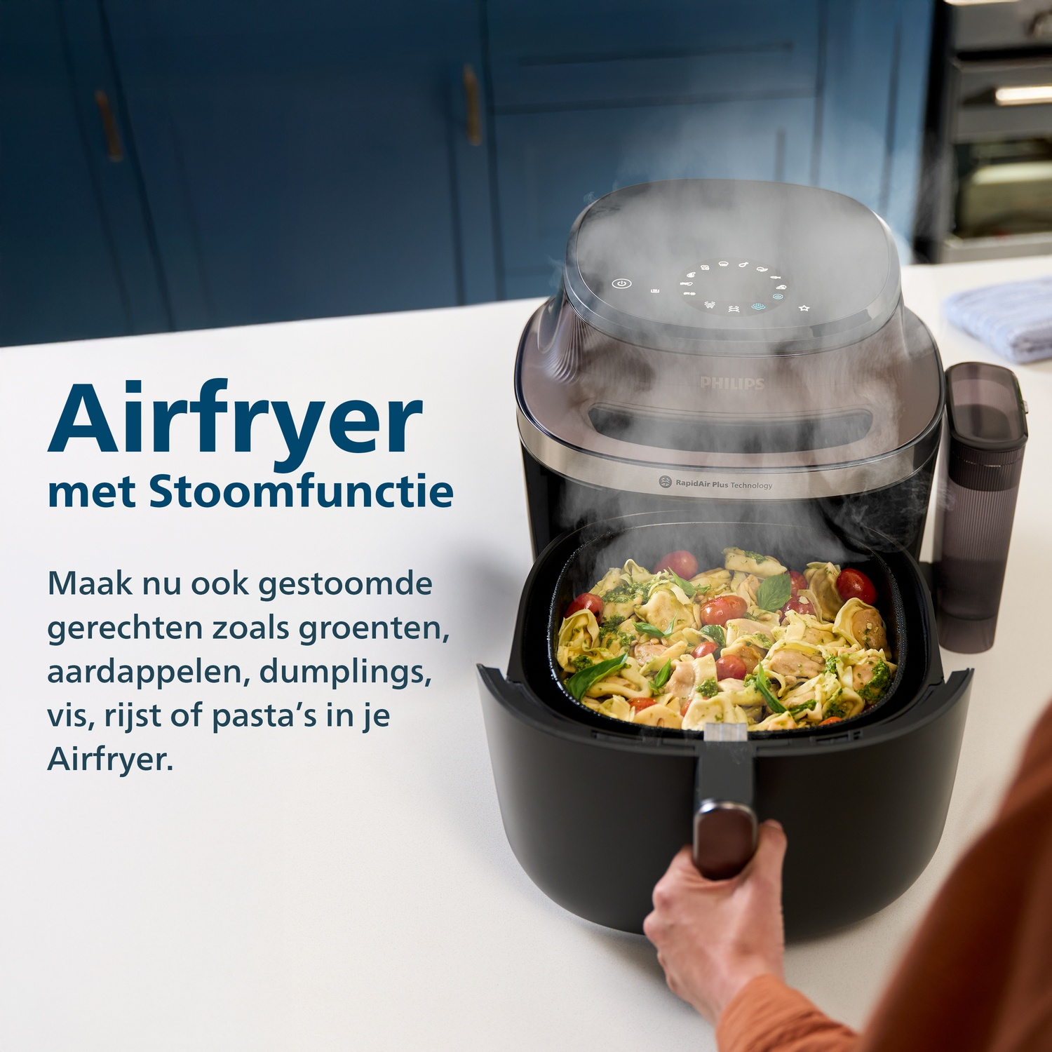 Een persoon houdt een airfryer met stoomfunctie vast, gevuld met pasta, tomaten en kruiden.