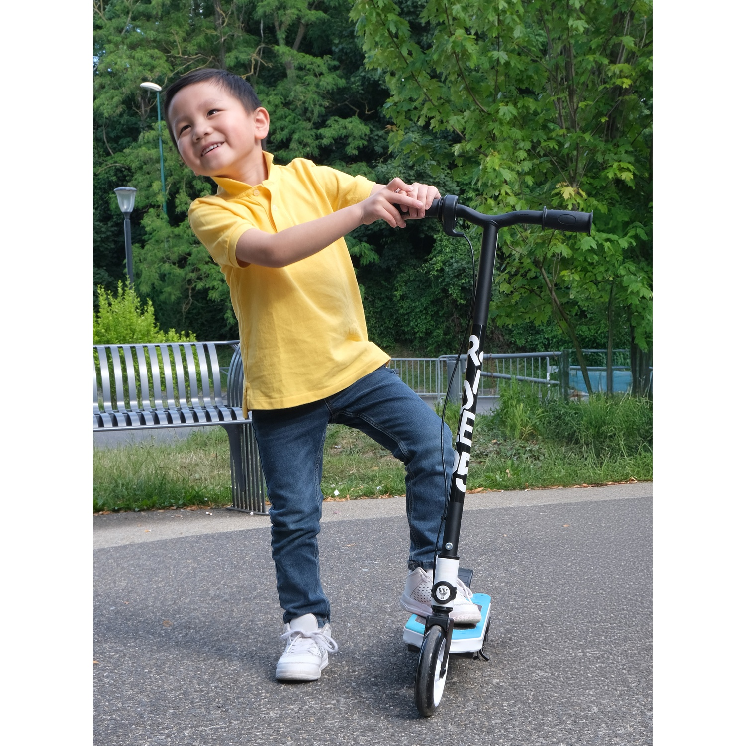 A boy in a yellow shirt and jeans stands on a scooter in a park.