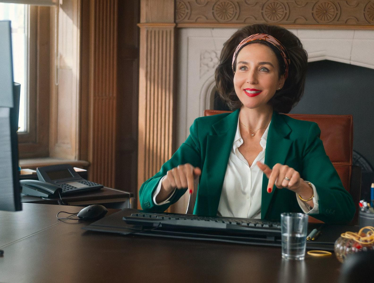 Eine Frau in einem grünen Blazer sitzt an einem Schreibtisch mit einem Computer.
