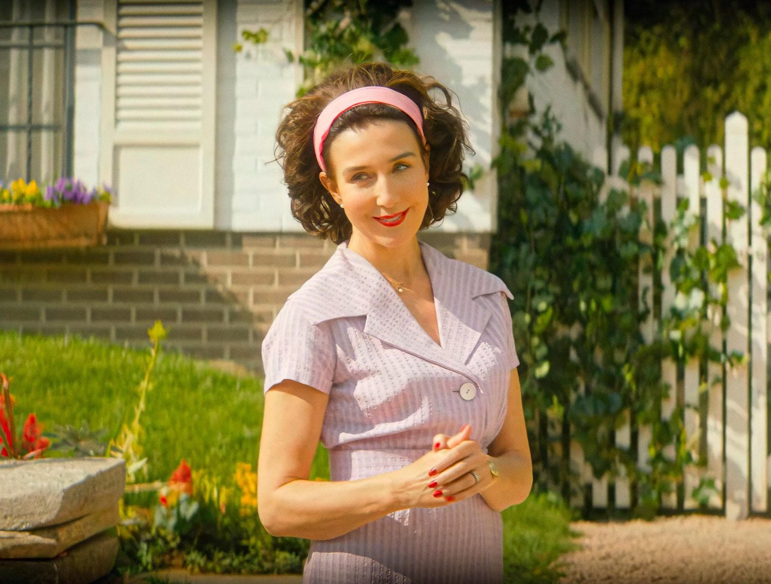 Frau mit rosa Stirnband und Kleid vor einem Haus mit Garten.