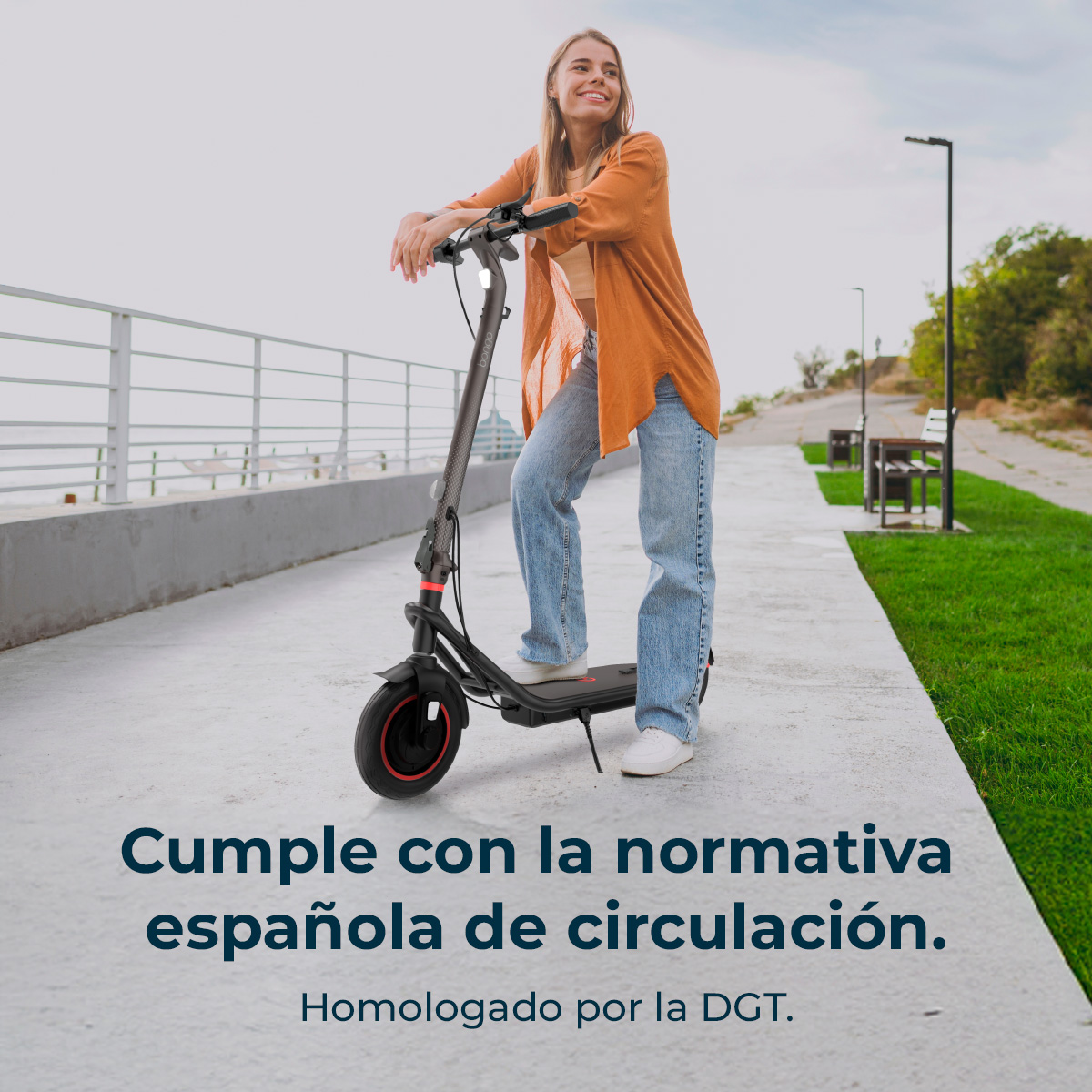 Una mujer está de pie en un patinete eléctrico en una acera. Lleva vaqueros y un cárdigan naranja.