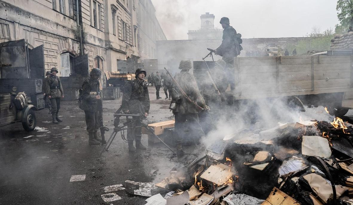 Soldaten in Uniformen stehen in einer rauchigen Gasse, Papiere brennen.