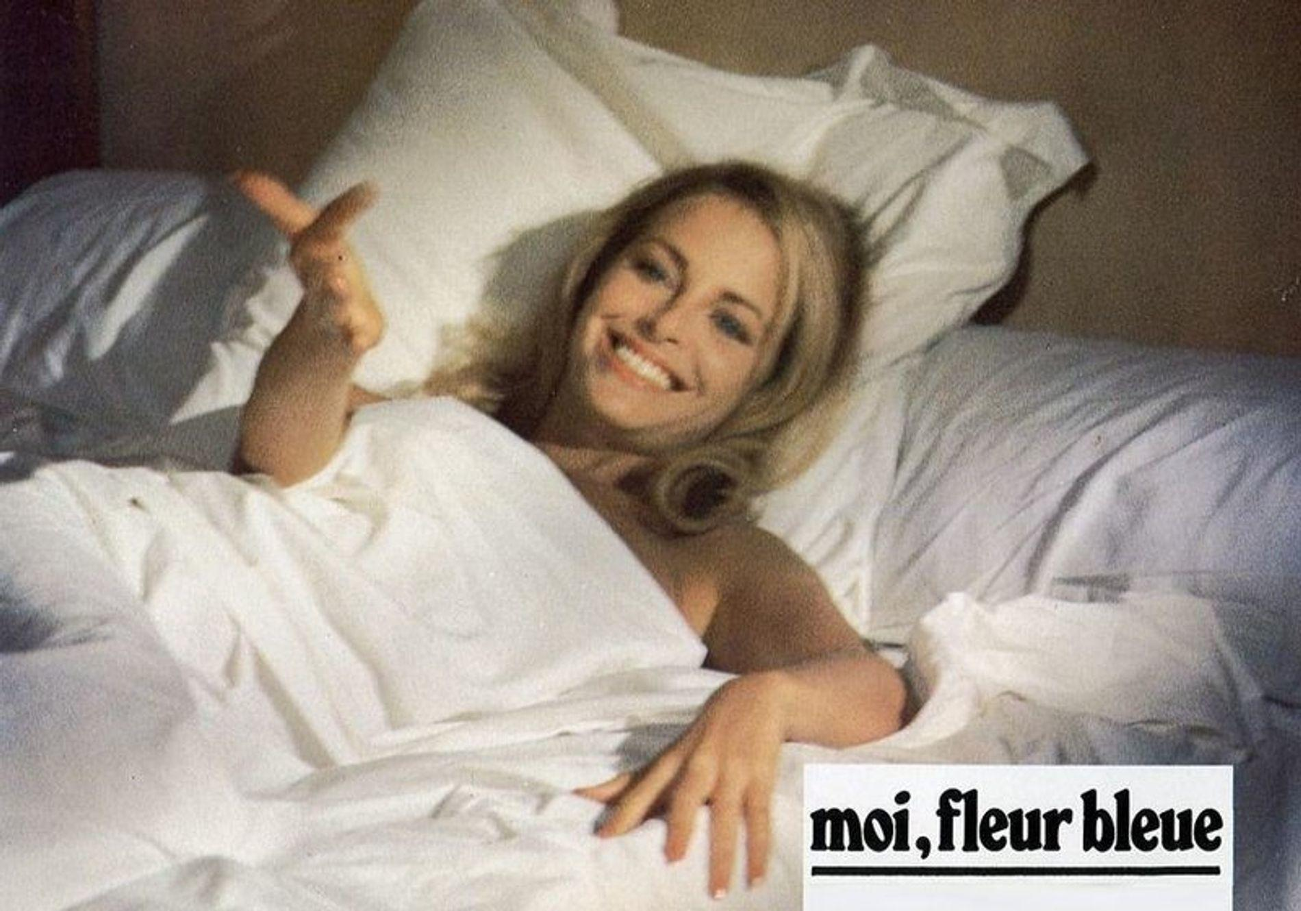 Eine lächelnde Frau mit blonden Haaren liegt in einem weißen Bett unter einer Decke.