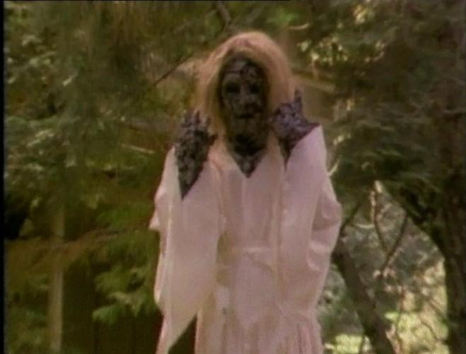 Eine verkleidete Person mit weißem Gewand und dunklem Gesicht steht im Wald.