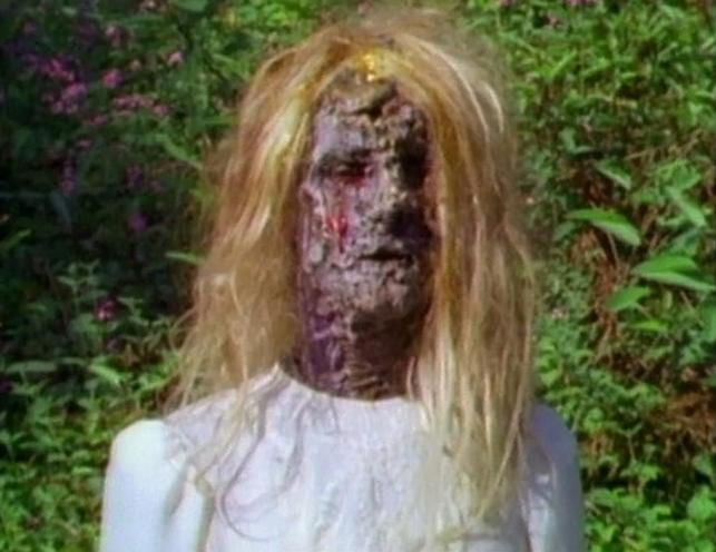 Eine verrottende Frau mit blonden Haaren und weißem Kleid steht im Freien.